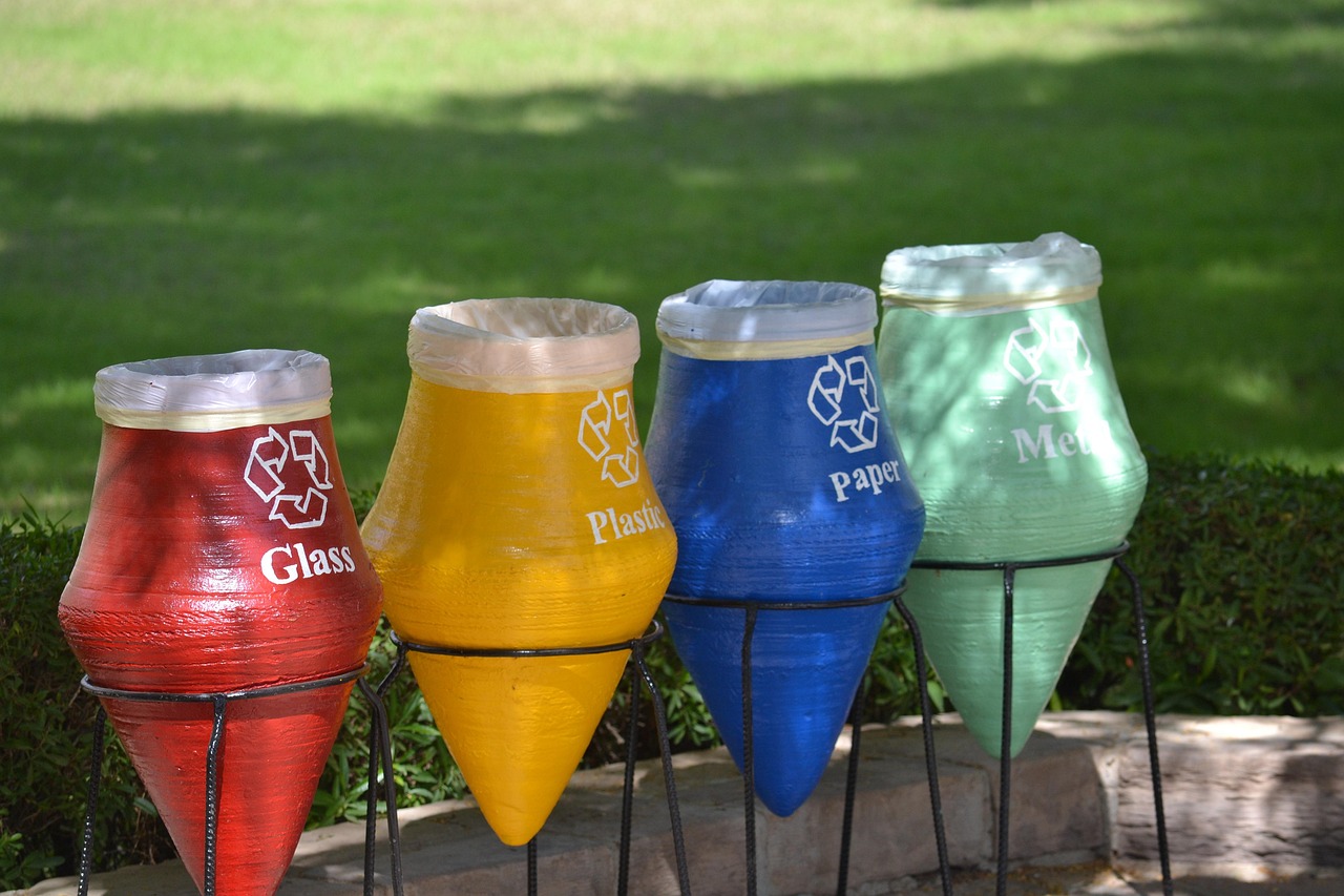 Quatro lixeiras coloridas de coleta seletiva sobre a grama: vermelha para vidro, amarela para plástico, azul para papel e verde para metal.